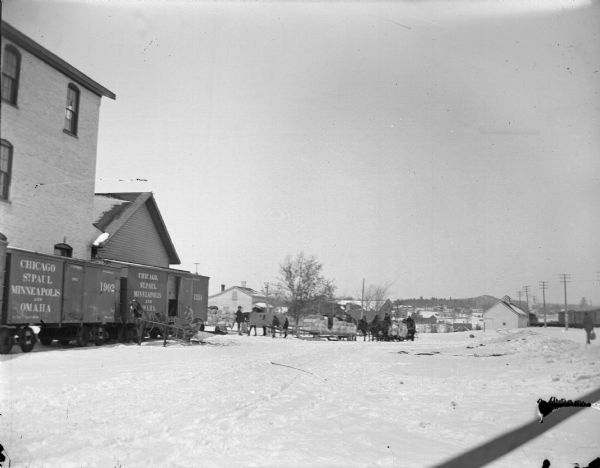 Loaded sleighs and wagons approaching the railroad station. Railroad cars are along the left.