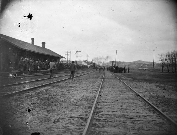 Train approaching railroad station crowded with people.