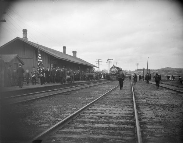Train approaching railroad station crowded with people and a band, possibly men coming or going from military service in World War I.