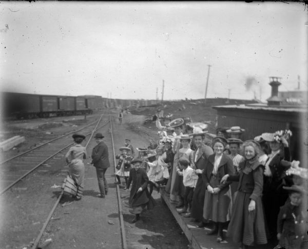 A crowd of happy, well-dressed adults and children wait at the railroad tracks for a train.