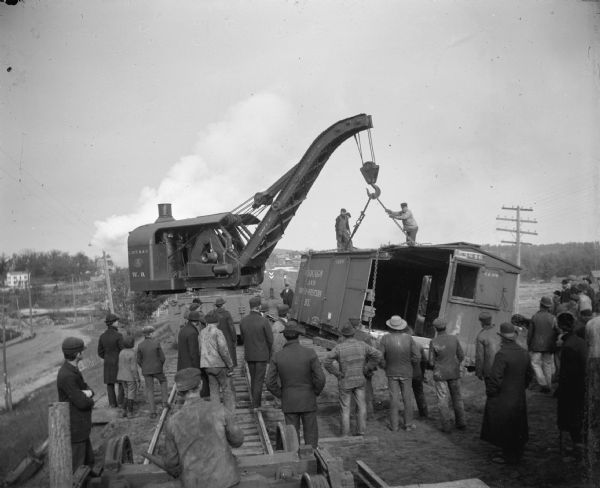 Crowd watching a railway crane lift a boxcar back on the track from the Chicago and Northwestern Railway.