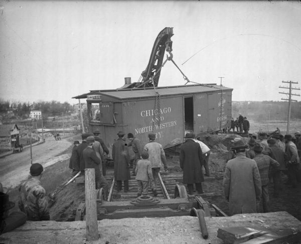 Crowd watching a railway crane lift a boxcar from the Chicago and Northwestern Railway back on the track.