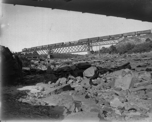Woman and child standing in the shadows among the rocks and logs along the river as a freight train crosses the elevated bridge above.