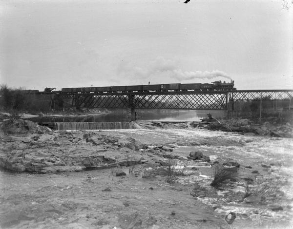 Men standing on top of a freight train crossing the river, and a log dam before the flood. Painted rocks in the river advertise for A.F. Werner.