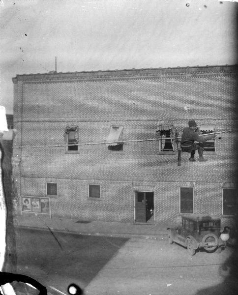 Elevated view towards a lineman sitting in a suspended seat, possibly stringing telephone line. There is an automobile parked near a brick building across the street.