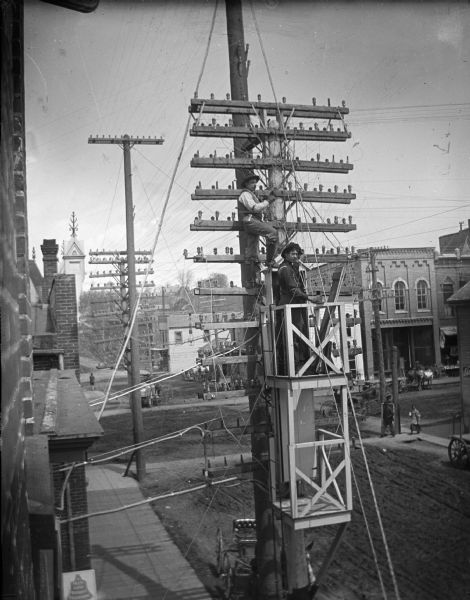 Elevated view of men working on telephone lines from scaffolding attached to the pole in front of the telephone exchange building.