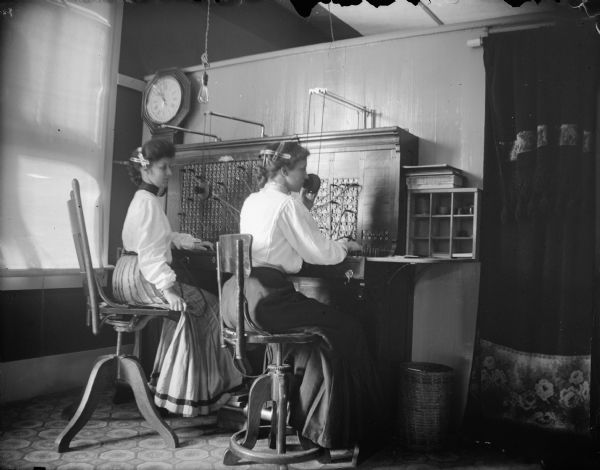 Two female telephone operators working at the telephone switchboard. Woman speaking into the mouthpiece of the switchboard.