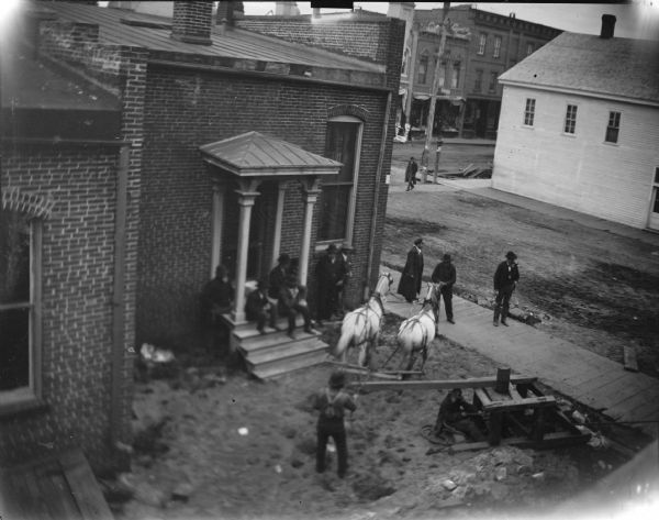 Elevated view of team of horses powering a winch for moving a house at the rear of the Jackson County Bank. Men are probably preparing the lot for the house.