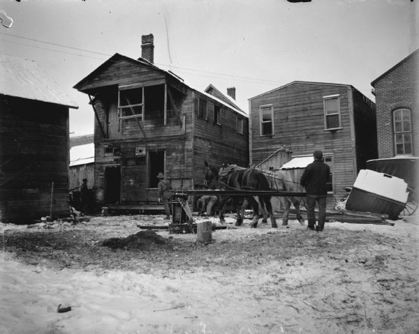 Team of horses powering a winch for moving a house. The winch was anchored to a post in the ground, logs were then placed under the house foundation and relocated as the structure moved, acting as rollers.