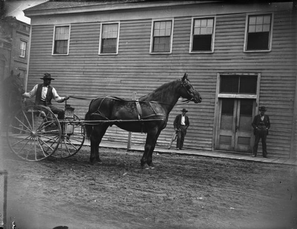 Man posing in a road cart, leading another horse. Two men stand on a board sidewalks in front of a large building on the other side of the street.