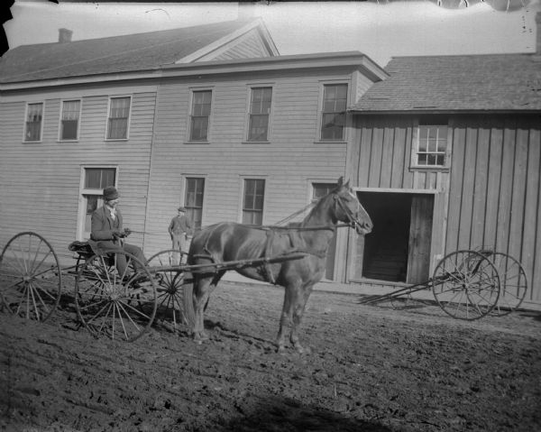 Man posing sitting on a light open buggy. The horse is harnessed in a way to permit speed. A man is standing on the board sidewalk in front of a large building on the other side of the street.