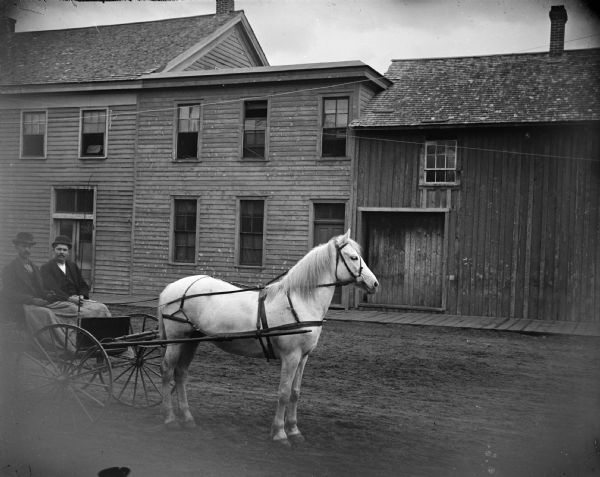 Two men in a wagon, pulled by a white horse, pause in the street.
