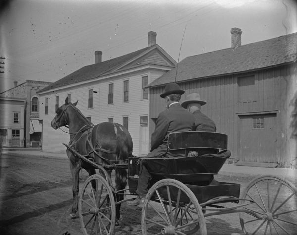 Rear view of horse and buggy pulling two men through town, with the man on the right possibly Bill Tester.