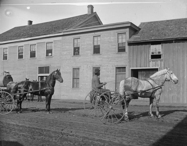 Man driving a road cart and horse. Another horse-drawn vehicle is on the left. Buildings are on the opposite side of the street.