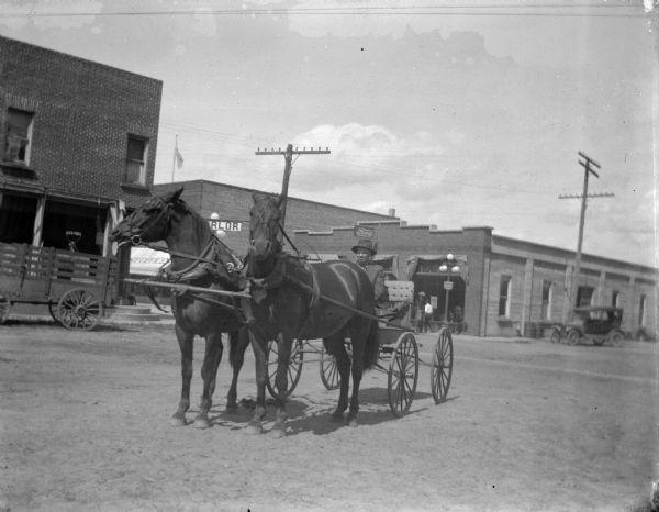 Man in a buggy driving a team of two horses through town, possibly Bill Testor who ran the Merchants Hotel. An automobile is visible in the right background, as well as a Ford showroom.