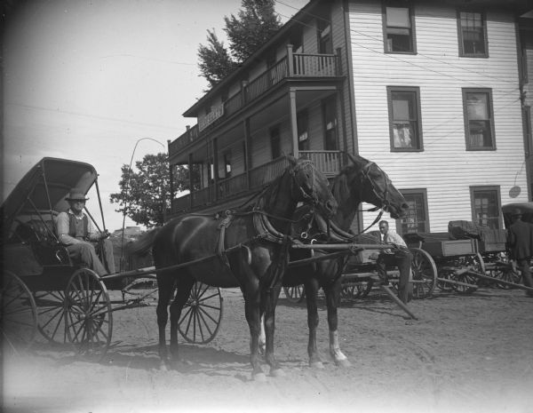 Man in a top-buggy posing with a team of two horses in front of the Merchants Hotel.