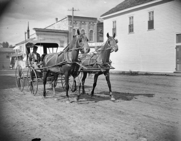 Man in a top-buggy driving a team of two horses through town.