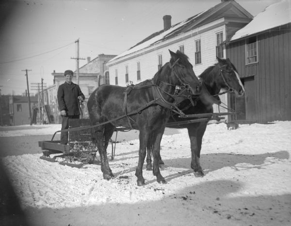 Man driving a bobsled and team of two horses. A row of buildings is along the street in the background.