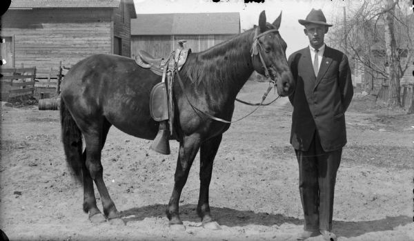 Man posing with a saddled horse. Farm buildings are in the background.