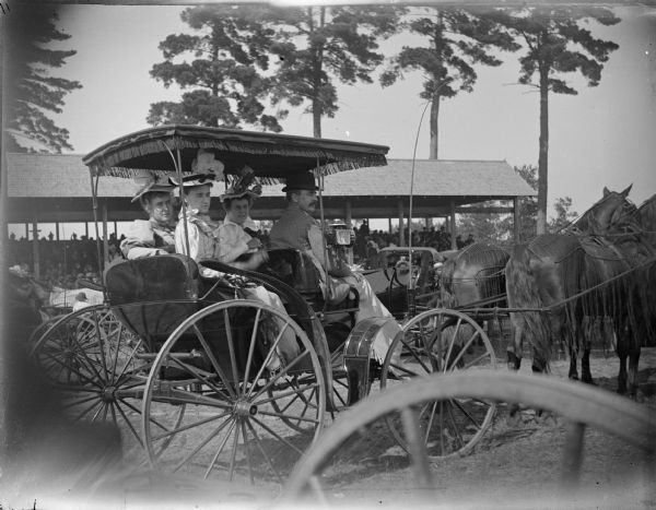 Three women and a man in a Spaulding surrey and two horse teams. Possibly the Spaulding family, and taken at the racetrack at the Jackson County Fairgrounds. The grandstand is in the background under tall trees.