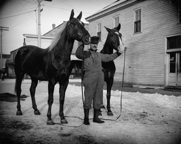 Man posing with two horses with white-blazed foreheads. Taken in town, with buildings in the background and snow on the ground.
