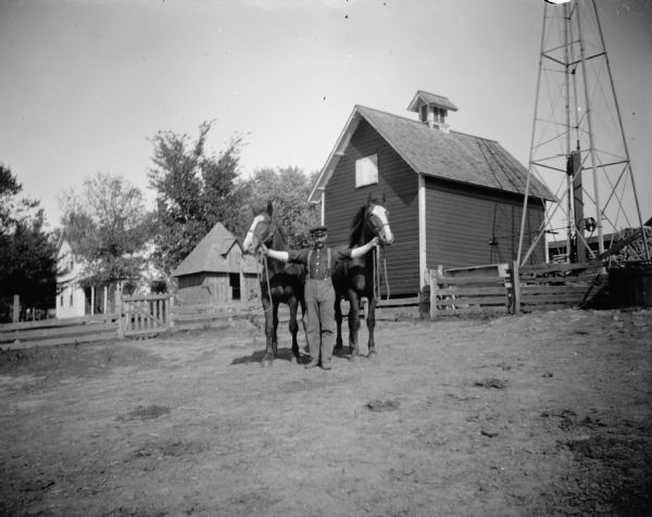 Man posing with two horses in a farmyard. Fences and farm buildings are in the background. A tower on the far right may be a windmill.