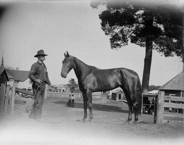 Man preparing to halter a race horse. They are standing near a fence, and in the background are buildings and people.