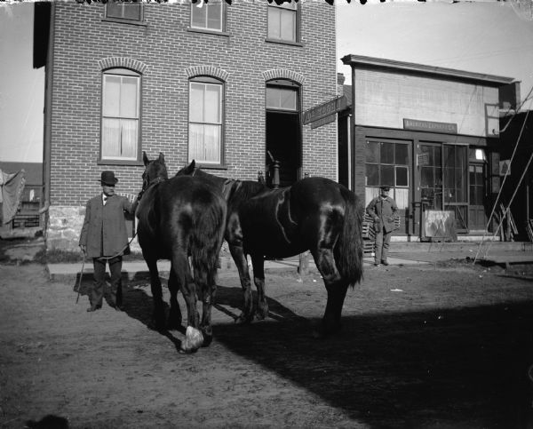 Man leading horses through town by the reins, in front of Dr. Krohn's Office and the Van Schaick Photograph Studio. Another man is standing in the background on the right.