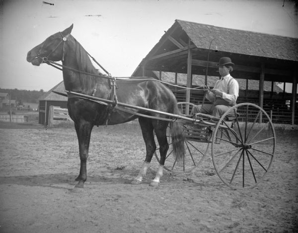 Man in a sulky posing at the racetrack, probably Moses Pauquette. There is an open-sided building in the background, perhaps a grandstand.