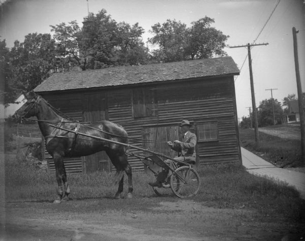 Man in a racing rig with a harnessed horse on the northwest corner of Harrison and Third Street. There is a barn behind them.