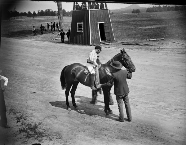 Elevated view from grandsstand towards men adjusting a jockey's stirrup and the gear of a horse on racetrack. On the other side of the track is the judges booth.
