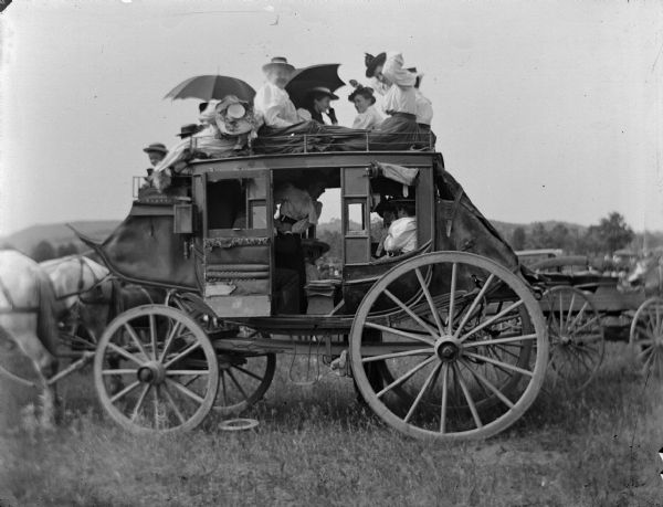 Women inside, and on top of a stagecoach with their umbrellas. A wagon is in the background on the right.