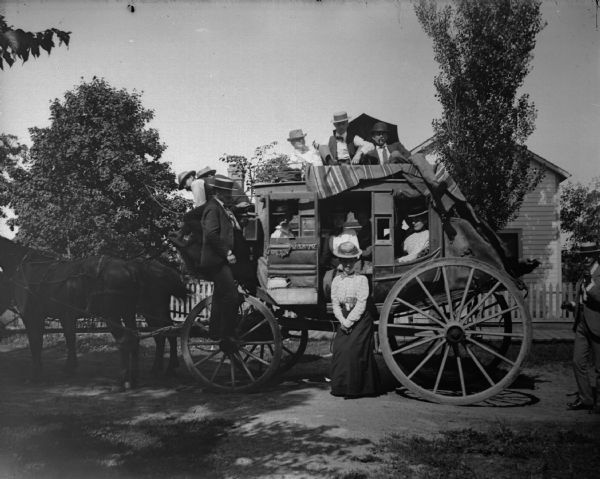 People crowding together on a stagecoach. It was kept in the Price barn and brought out for special occasions like the Jackson County Fair. Passengers were considered to be the elite of Black River Falls. The driver was Ed Pratt, a highly respected associate of W.T. Price. W.T. Price had operated a stage line from Sparta via Black River Falls to Eau Claire via Hixton and Osseo. When the railroad was built the stage line was discontinued. A house is in the background behind trees.