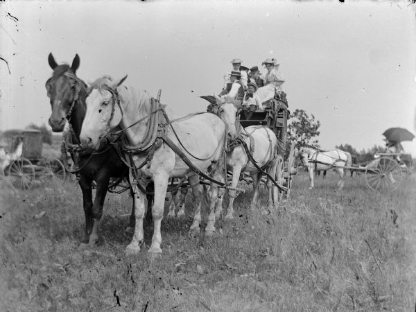 Group of people posing on top of stagecoach. It was a special occasion when the old stagecoach was brought out. The top-loading was probably done to get everyone in the photograph. Other horse-drawn vehicles are in the field in the background.