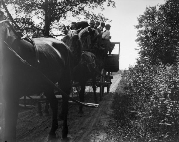 Group of people riding on a stagecoach looking back down the road. The horse pulling the stagecoach is in the foreground.
