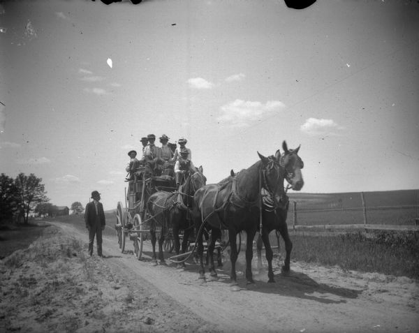 Group on a stagecoach pulled by a team of four horses. A man is standing on left in the road next to the stagecoach. In the left background are buildings and trees.