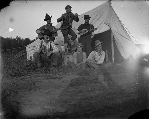 Five men and a boy outside a tent, one dancing with snakes, two others playing a guitar and mandolin.