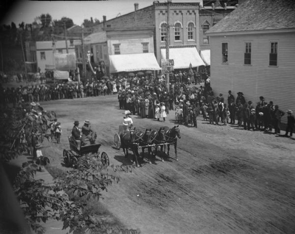 Elevated view of a crowd watching a woman in a chariot driving four horses in a circus parade in town.