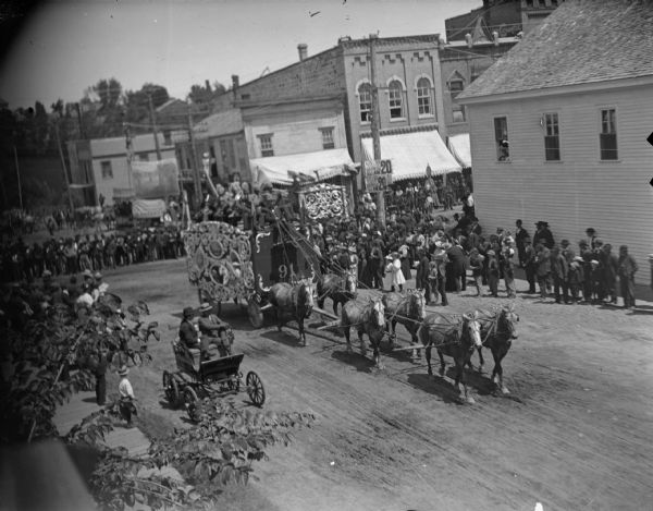 Elevated view of a crowd watching a circus wagon pulled by a team of six horses in a circus parade in town.