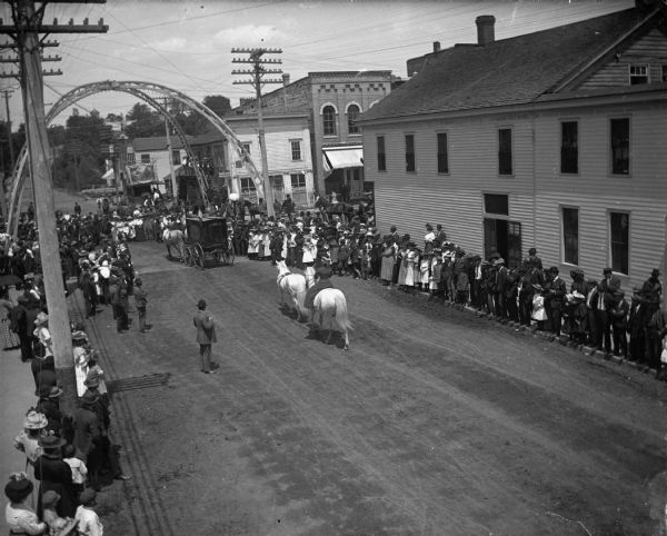 Elevated view of a parade passing under arch at the intersection of First and Main Streets. Crowds of people are watching from the sidewalks.