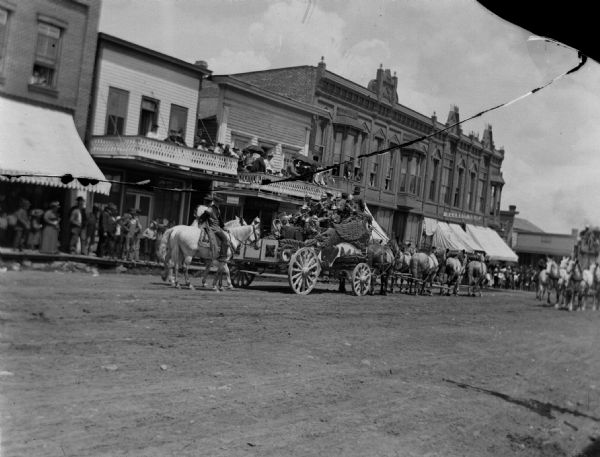 View across street towards a band in a wagon parading through town. People are watching from the sidewalk, and also from second story porches.