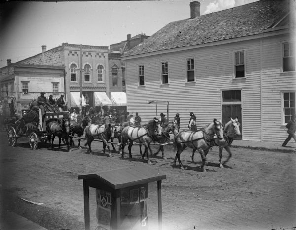 Slightly elevated view over sidewalk towards eight horses drawing a circus wagon in parade. The parade is coming from the circus ground at the white pine grove in the bend of the river, going north on First Street. Andrew Froen's Saloon is on the right, and Charles J. Van Schaick's Photography Gallery showcase is at the bottom center.