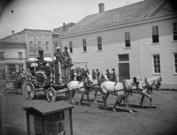 Slightly elevated view of men driving a circus calliope for a parade in town. A calliope is a musical instrument that produces sound by sending steam through whistles, originally locomotive whistles. Charles J. Van Schaick's Photography Gallery showcase is at the bottom center.
	