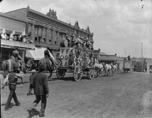 Rear view of a circus wagon going through town, with a band on top pulled by a team of six horses. People are walking in the street behind the wagon, and two people are on horseback. Across the street people are watching from the sidewalk, and from second-story porches.
