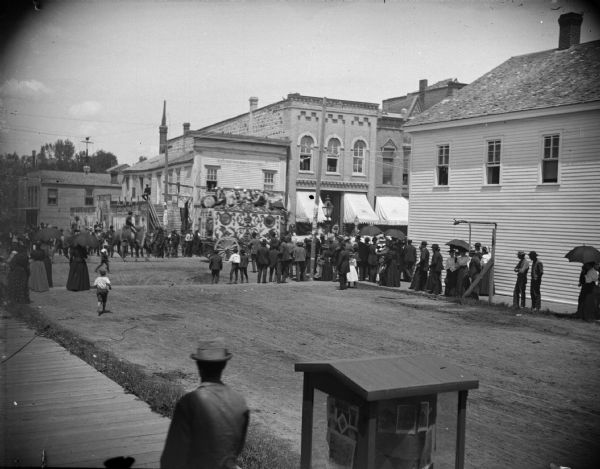 Crowd watching a circus wagon with a band on top, in a parade in town, followed by a camel.	