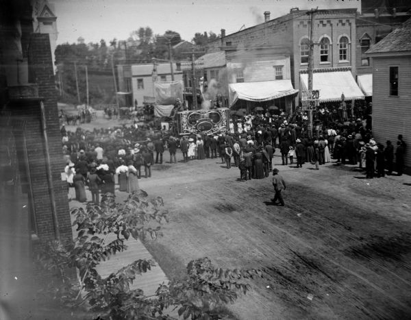 Elevated view towards intersection of a crowd watching a circus wagon on Main Street, probably a steam calliope.
