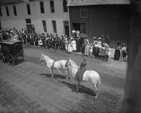 Elevated view of a crowd watching a woman with two white horses in a circus parade. She is riding one of the horses.