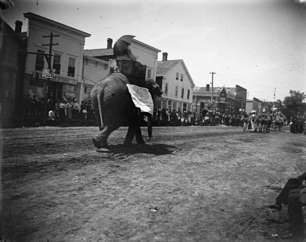 Rear view of a man riding an elephant in a circus parade through town. Crowds line the sidewalk on the other side of the street.