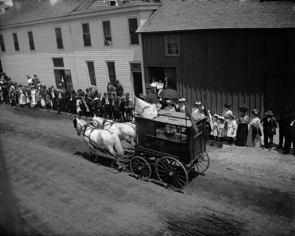 Elevated view of a man in a parade driving a circus wagon with a cage holding a leopard. A crowd lines the sidewalk on the other side of the street.