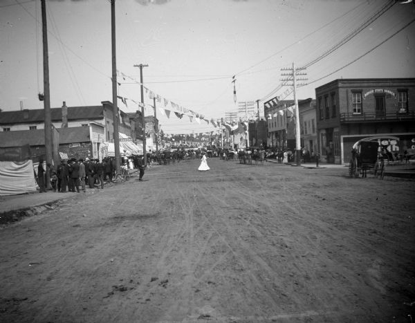 View down center of unpaved street towards a crowd gathering on patriotically decorated Main Street. The <i>Badger State Banner</i> is at the left, the home of the local newspaper.